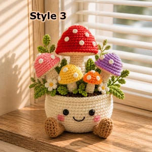 May include: A crochet arrangement featuring a cream-colored pot with a smiling face and two brown feet, containing a cluster of colorful mushrooms in red, pink, yellow, orange, and purple, accented with small white flowers and green leaves.