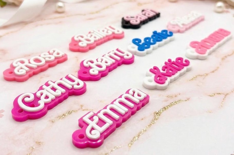 Może przedstawiać: Kolekcja spersonalizowanych zawieszek z imionami w r&oacute;żnych kolorach. Zawieszki przedstawiają imiona takie jak "Cathy", "Emma" i "Katie" w białych literach na r&oacute;żowym, niebieskim i czarnym tle. Zawieszki są ułożone na marmurowej powierzchni.
