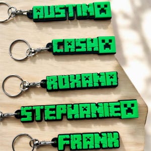May include: Keychains with names in a pixelated font. The names "AUSTIN", "CASH", "ROXANA", "STEPHANIE", and "FRANK" are displayed in green letters with black outlines. Each keychain has a silver-colored ring and clasp.