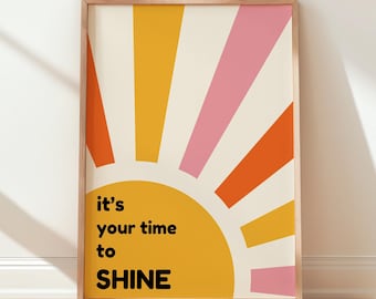May include: A framed art print featuring a stylized sunburst design in yellow, orange, and pink hues against a cream background. The text "it's your time to SHINE" is displayed in black. The artwork is in a wooden frame.