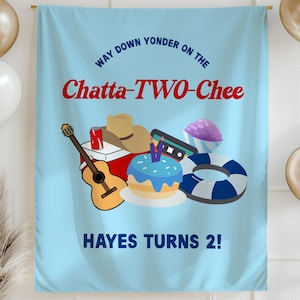 May include: A light blue banner with the text "Way down yonder on the Chatta-TWO-Chee" and "Hayes turns 2!". The banner features illustrations of a guitar, cake, hat, and other objects. Gold and white balloons are in the background.
