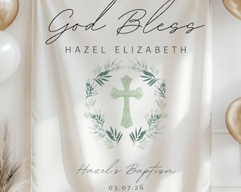 May include: A white banner with the text "God Bless" and "Hazel Elizabeth" in elegant script. A green cross is centered within a wreath of leaves. The banner also reads "Hazel's Baptism" and the date 03.07.26. Gold and white balloons are on either side.