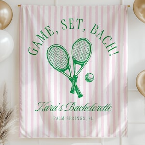 May include: A pink and white striped banner with the text "GAME, SET, BACH!" and tennis rackets. The banner also includes the text "Kara's Bachelorette" and "PALM SPRINGS, FL". Gold and white balloons are on either side.