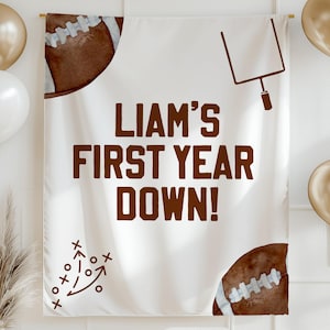 May include: A white banner with the text "LIAM'S FIRST YEAR DOWN!" in brown. The banner features football graphics and a goalpost illustration. Gold and white balloons and decorative elements complete the festive scene.