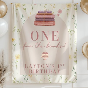 May include: A cream-colored birthday banner with floral and book illustrations. The banner reads "ONE for the books!" and "LAYTON'S 1ST BIRTHDAY". Gold and white balloons and a gold banner are also in the image.