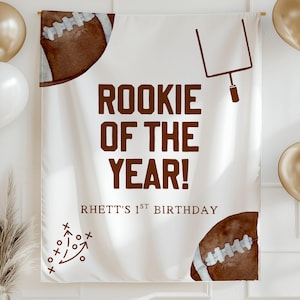Könnte beinhalten: Ein weißes Banner mit dem Text "ROOKIE OF THE YEAR!" in Braun. Das Banner hat Football-Grafiken und den Text "RHETT'S 1ST BIRTHDAY". Goldene und weiße Luftballons und eine dekorative Pflanze sind im Bild.