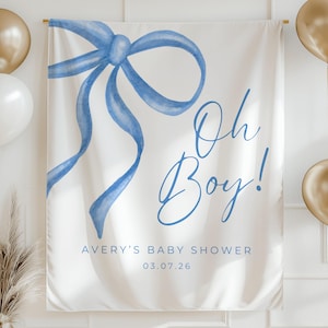 Può includere: Un banner bianco per baby shower con un fiocco blu ad acquerello e la scritta "Oh Boy!". Il banner include anche il testo "Avery's Baby Shower" e la data 03.07.26. Palloncini dorati e bianchi su entrambi i lati.