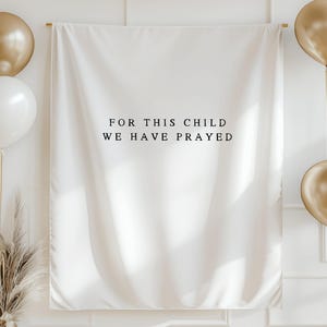 May include: A white backdrop with the text "FOR THIS CHILD WE HAVE PRAYED" in black. The backdrop is decorated with gold balloons, a gold banner, and a gold vase with dried plants. The scene is set against a white wall.