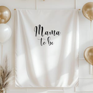 May include: A white fabric backdrop with the words "Mama to be" in black script, gold and white balloons, and gold triangle bunting. A gold vase with dried plants is in the lower left corner. The backdrop is hung on a wooden rod.