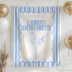 May include: A cream-colored banner with blue vertical stripes and text that reads "Welcome to Lauren's Bachelorette" and "Rosemary Beach." A drawing of a woman in a martini glass is also featured. Gold and white balloons and gold triangle bunting decorate the scene.