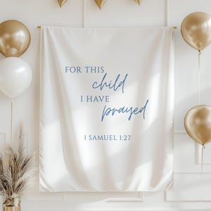 May include: A white banner with blue script reading "FOR THIS child I HAVE prayed 1 SAMUEL 1:27". The banner is suspended from a wooden rod, with gold and white balloons. Gold triangular bunting is suspended above.