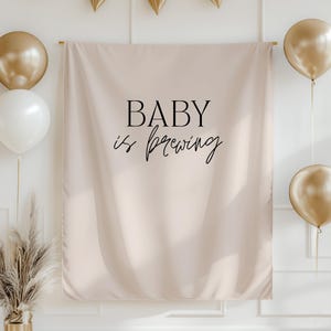 May include: A beige backdrop with the text "BABY is brewing" in black script, decorated with gold balloons and a gold banner. A gold vase with dried pampas grass is in the lower left corner. The scene is set against a white wall.