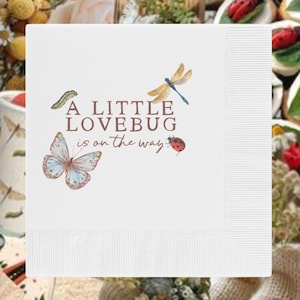 May include: A square white paper napkin featuring a brown printed text that reads A LITTLE LOVEBUG is on the way. The design includes a green caterpillar, a blue and white butterfly, a brown dragonfly, and a small red ladybug with black spots.