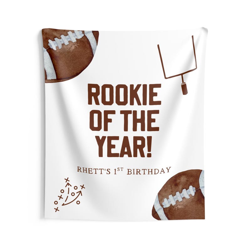 May include: White fabric wall hanging with brown text that reads "Rookie of the Year!" and "Rhett's 1st Birthday". The design includes watercolor footballs, a football play diagram, and a goalpost.