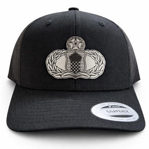 May include: Black trucker hat with a silver emblem featuring a shield, star, and laurel wreath design. The hat has a curved brim and a mesh back. The hat has a snapback closure.