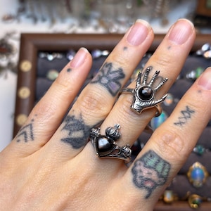 May include: Two silver rings are shown on a hand. One ring has a hand design with a dark stone, and the other is a heart-shaped ring with a crown design and a dark stone. Other jewellery is visible in the background.