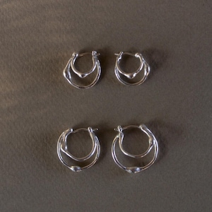 Puede incluir: Cuatro pares de pendientes de aro plateados. Cada pendiente presenta un diseño de doble aro con un pequeño detalle redondeado en la parte inferior. Los pendientes se muestran sobre un fondo marrón neutro, resaltando su estilo minimalista.