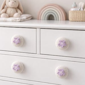 May include: White chest of drawers with four crocheted drawer knobs. Each knob has a light purple flower design. The chest is in a nursery setting, with a stuffed bunny and wooden rainbow on top.