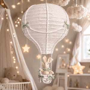 May include: A white paper hot air balloon mobile featuring a ribbed texture and sage green ribbon bows. A small plush rabbit sits inside the attached white basket, which is also decorated with matching sage green ribbon bows on the sides.