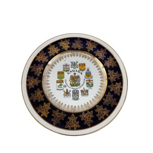 May include: A white ceramic plate with a blue and gold border. The center of the plate features a design of the coats of arms of the provinces and territories of Canada. The text "Canada Coats of Arms & Emblems" is printed on the plate.