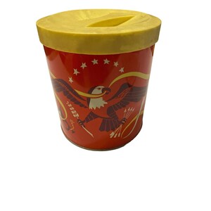 Puede incluir: Lata de metal roja con tapa amarilla y un gráfico de un águila calva con estrellas y rayas. La lata tiene una ranura en la tapa.