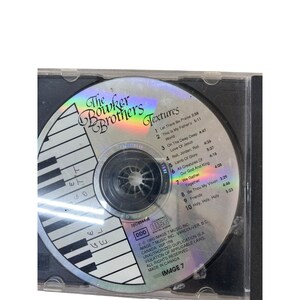 May include: A silver compact disc with a black and white keyboard design. The disc is labeled "The Bowker Brothers Textures" and lists the song titles and lengths. The disc is in a clear plastic case.