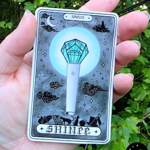 May include: A rectangular sticker featuring a stylized light blue diamond-shaped object on a white handle, set against a backdrop of a full moon and stars. The sticker has a gray border with the word "SHINee" at the bottom and "MMVIII" at the top.
