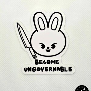 May include: A white sticker featuring a cartoon rabbit head with a knife and the words "BECOME UNGOVERNABLE" in black. The rabbit has a stern expression. A small logo with a moon and stars is in the bottom right corner.
