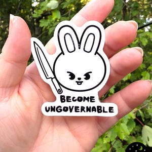 May include: A white sticker featuring a cartoon bunny holding a knife. The bunny has a simple design with black outlines and a small, mischievous expression. The text "BECOME UNGOVERNABLE" is below the bunny.