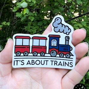 May include: A sticker featuring a cartoon train with red and blue cars, a blue engine, and a white smoke cloud. The text "IT'S ABOUT TRAINS" is printed in black on a white background.