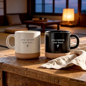 May include: Two ceramic mugs, one white and one black, both featuring a speckled finish and a solid tan base. Each mug displays the printed text 'you custom text here' above a small hand-drawn heart icon. The mugs have a smooth, rounded handle.