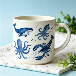 May include: A cream-coloured ceramic mug featuring a blue painted pattern of various sea creatures including a whale, an octopus, a squid, and a lobster. The mug has a smooth finish, a rounded handle, and a cylindrical shape with a wide opening.