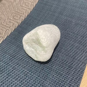 Puede incluir: Una piedra lisa de color verde claro, de forma redondeada. Descansa sobre un mantel individual azul oscuro tejido. La piedra tiene un aspecto translúcido, lo que sugiere que es natural y sin pulir.