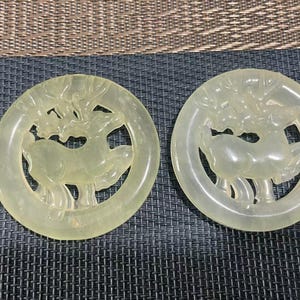May include: Two pale yellow jade pendants. Each pendant features a deer design within a circular frame. The deer is depicted with antlers and a stylized body. The pendants are displayed on a dark gray textured surface.