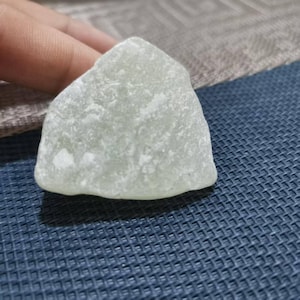 Puede incluir: Una piedra verde pálido de forma irregular con una superficie texturizada. La piedra tiene un aspecto ligeramente translúcido y una base redondeada, con una parte superior más puntiaguda. La piedra es probablemente un objeto decorativo o un espécimen natural.