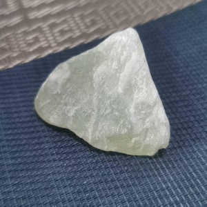 Puede incluir: Una gema verde pálido de forma irregular, con una superficie rugosa y texturizada. La piedra es translúcida y parece ser un espécimen mineral natural. Fondo azul oscuro texturizado.