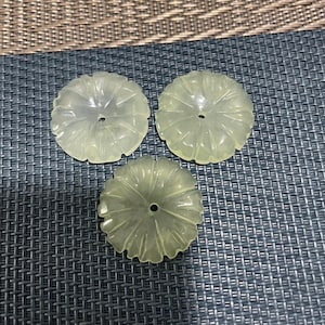 May include: Three translucent, pale green, flower-shaped jade pendants. Each pendant has a central hole and a ribbed design radiating from the center. The pendants are displayed on a textured, dark gray surface.