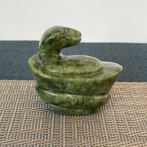 May include: A carved green stone trinket box shaped like a coiled snake. The lid features a sculpted snake head with visible eyes and scales, resting on a rounded base. The stone has a mottled dark and light green appearance with a polished finish.
