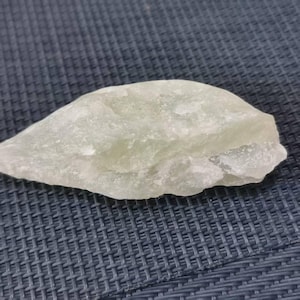 Puede incluir: Una piedra de color verde pálido, de forma irregular y con una superficie rugosa y texturizada. La piedra tiene una cualidad translúcida, y su forma es orgánica y natural. La roca se encuentra sobre un fondo azul oscuro tejido.