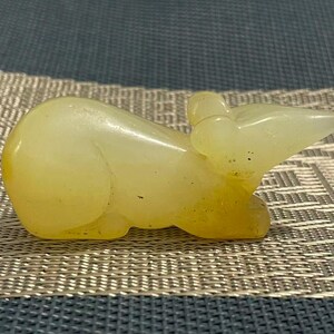Op de afbeelding: Een klein, glad, lichtgeel uitgesneden stenen beeldje in de vorm van een muis. Het beeldje heeft een afgerond lichaam, een spitse snuit en kleine oortjes, met een doorschijnende kwaliteit en subtiele goudkleurige variaties in het materiaal.