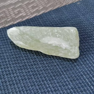 Puede incluir: Una piedra verde pálido de forma irregular con una superficie lisa y pulida. La piedra tiene una cualidad ligeramente translúcida y descansa sobre una superficie texturizada azul oscuro. La forma de la piedra es orgánica y natural.