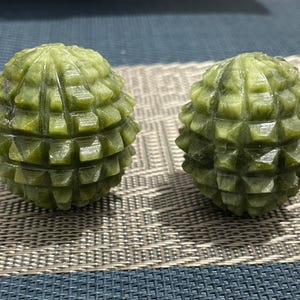Peut inclure: Deux sphères en pierre verte sculptée avec un motif géométrique texturé. Les sphères ont une apparence épineuse et segmentée et sont exposées sur une surface à motifs. La couleur est un mélange de vert clair et foncé.