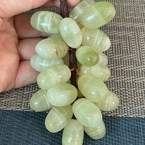 May include: A decorative cluster of light green, translucent stone grapes with visible white banding. The individual oval-shaped stones are attached to a central dark brown, wrapped wire stem, creating a realistic bunch of grapes sculpture.