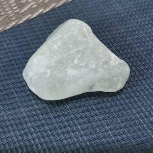 Puede incluir: Una piedra lisa y redondeada, de color verde claro, con una forma ligeramente triangular. La piedra tiene un aspecto esmerilado y está sobre una superficie texturizada de color azul oscuro. El color de la piedra es un verde pálido y apagado.