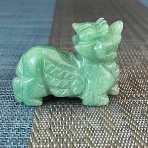 May include: A small, light green carved stone figurine depicting a mythical creature with wings, a prominent head crest, and a textured body. The sculpture features smooth, polished surfaces and detailed etchings that define the wings and facial features.