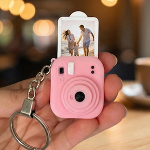 May include: A pink miniature camera keyring displaying a small photo. The camera features a black lens and a silver key ring. The photo shows a couple on a beach. The background includes a blurred coffee cup and bokeh lights.
