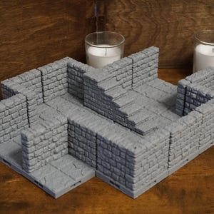 May include: A modular gray dungeon terrain set featuring interlocking stone walls and a staircase section. The pieces are constructed with a textured brick pattern, designed to be arranged into various room configurations for tabletop gaming.