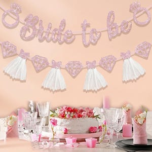 May include: A pink glitter banner that reads "Bride to be" with diamond ring and dress accents. The banner is strung above a table set for a celebration, including a cake, candles, and glassware. The overall theme is a bridal shower.