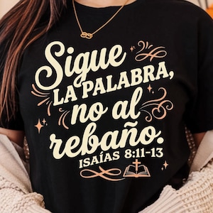 May include: Black t-shirt with the Spanish text "Sigue la palabra, no al rebaño" in cream and peach colors. The text includes the Bible verse "Isaías 8:11-13" and a small illustration of an open book and cross. The shirt has rolled-up sleeves.