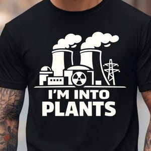 以下が含まれることがあります： 白いグラフィックの発電所と「I'M INTO PLANTS」の文字がプリントされた黒いTシャツ。デザインには煙突、建物、送電塔が含まれています。Tシャツはクルーネックです。